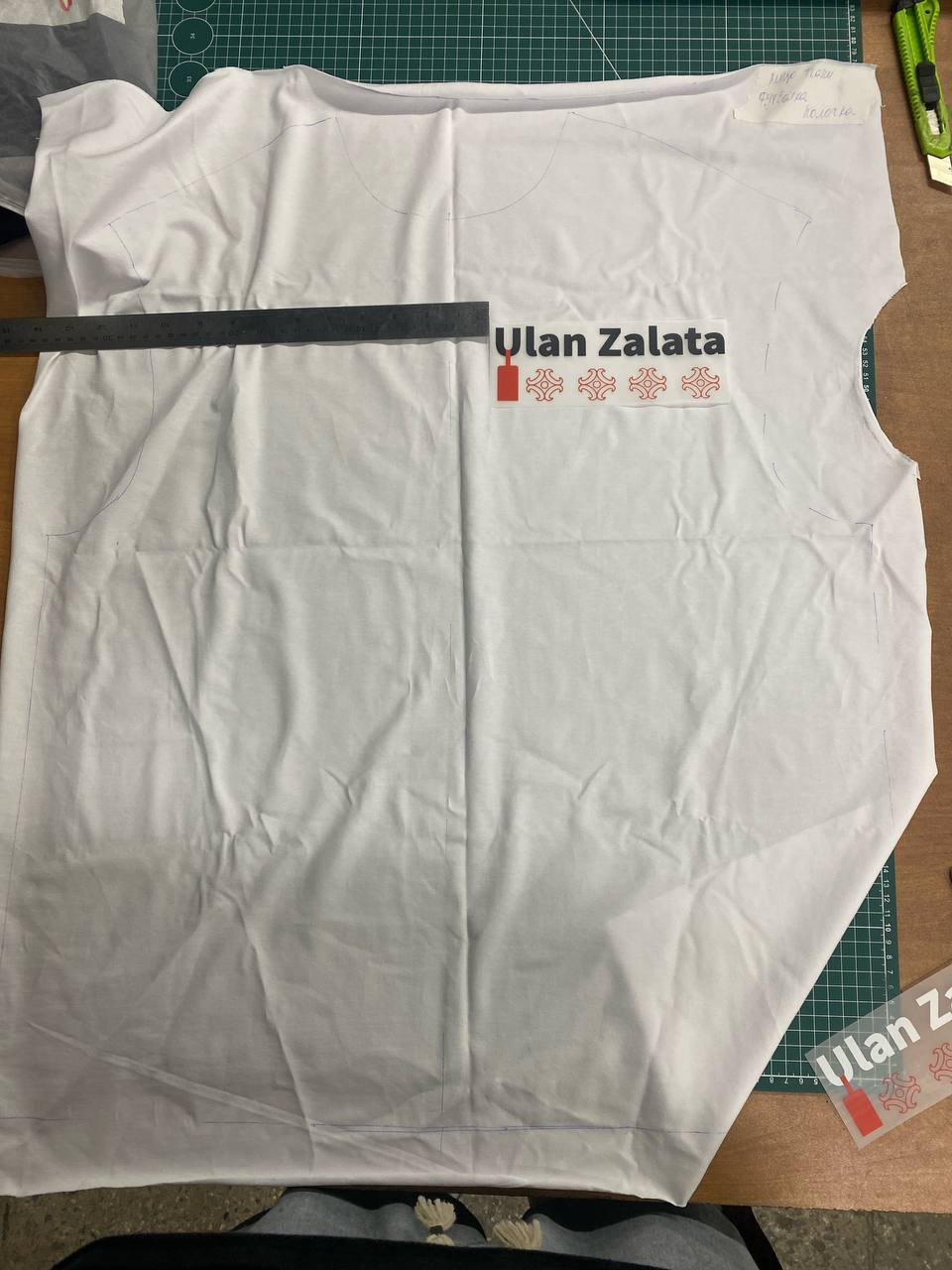Раскроенная белая футболка на столе: логотип Ulan Zalata примеряют по линейке перед нанесением